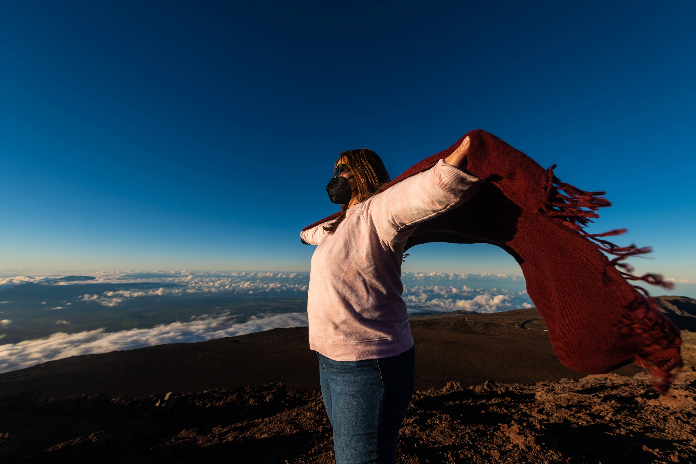 Female wearing pink sweater with her arms spread wide with a red cotton scarf standing on top a hill at Haleakala National Park, Maui, Hawaii