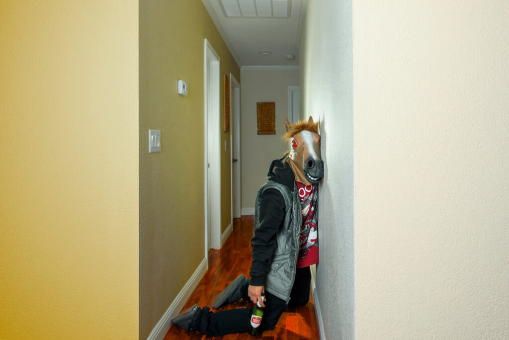 Person with horsehead mask on leaning drunk against the wall holding a green beer bottle in one hand.