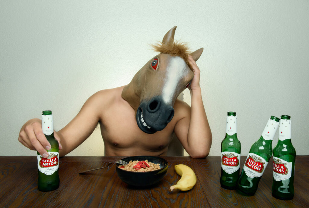 Shirtless Person with Horsehead Mask on head with one hand holding his head and the other holding a green beer bottle. A bowl of cereal and a yellow banana on a table with three additional, empty green beer bottles leaning in an abnormal fashion and a fork resting on edge of cereal bowl - The Day After Series (Conceptual Work)