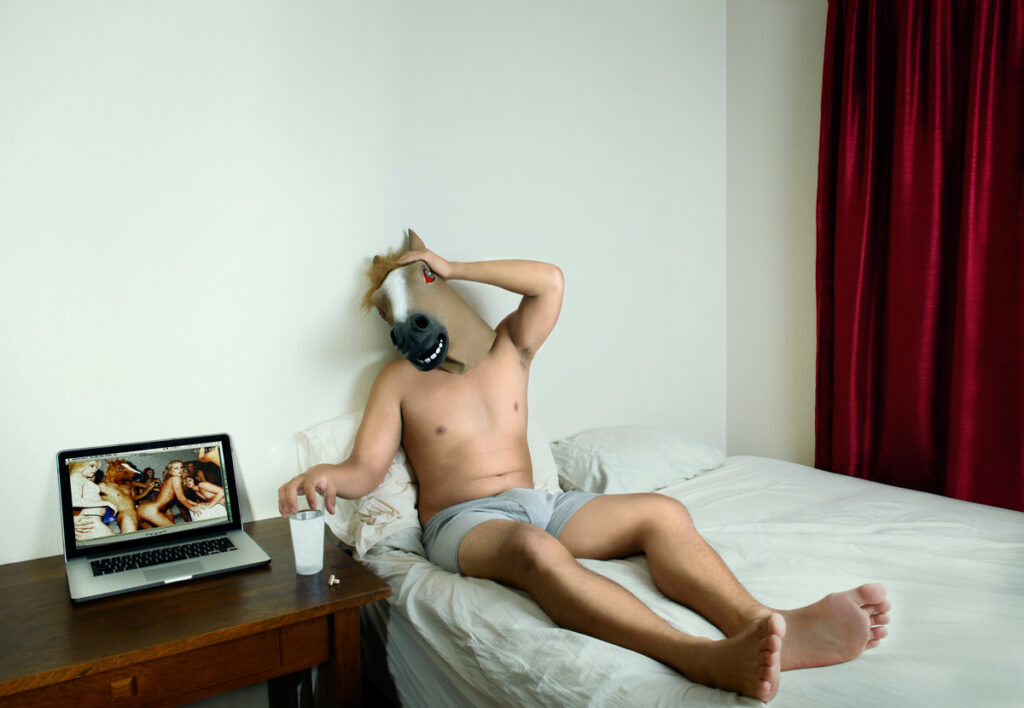 Man with Horsehead mask on a bed with only his briefs on,holding one hand behind head with the other on a cup of water that sits next to a laptop replaying the scene that went on the night before - Final Image for 'The Day After Series'(Conceptual Work)