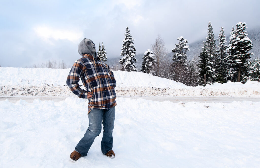 Alien-Masked person wearing long blue jeans and blue and orange long sleeve plaid shirt standing on white snow with both hand behind hip, arching back and looking up to the sky. Hill of sand in the background with four pine trees in the distant with sections of white snow - 'Alienated' Series
