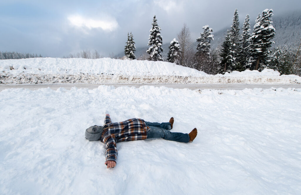Alien-Masked person wearing blue jeans and orange and blue long sleeve plaid shirt with both arms spread wide open laying on white snow. In the distant, there are pine trees with sections of snow on top of leaves - 'Alienated' Series
