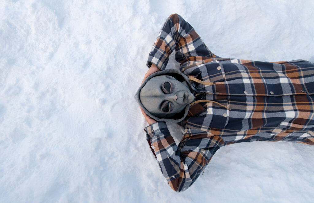 Alien-Masked person wearing a blue,orange plad longsleeve shirt laying on white snow with both arms raised above and behind head - 'Alienated' Series