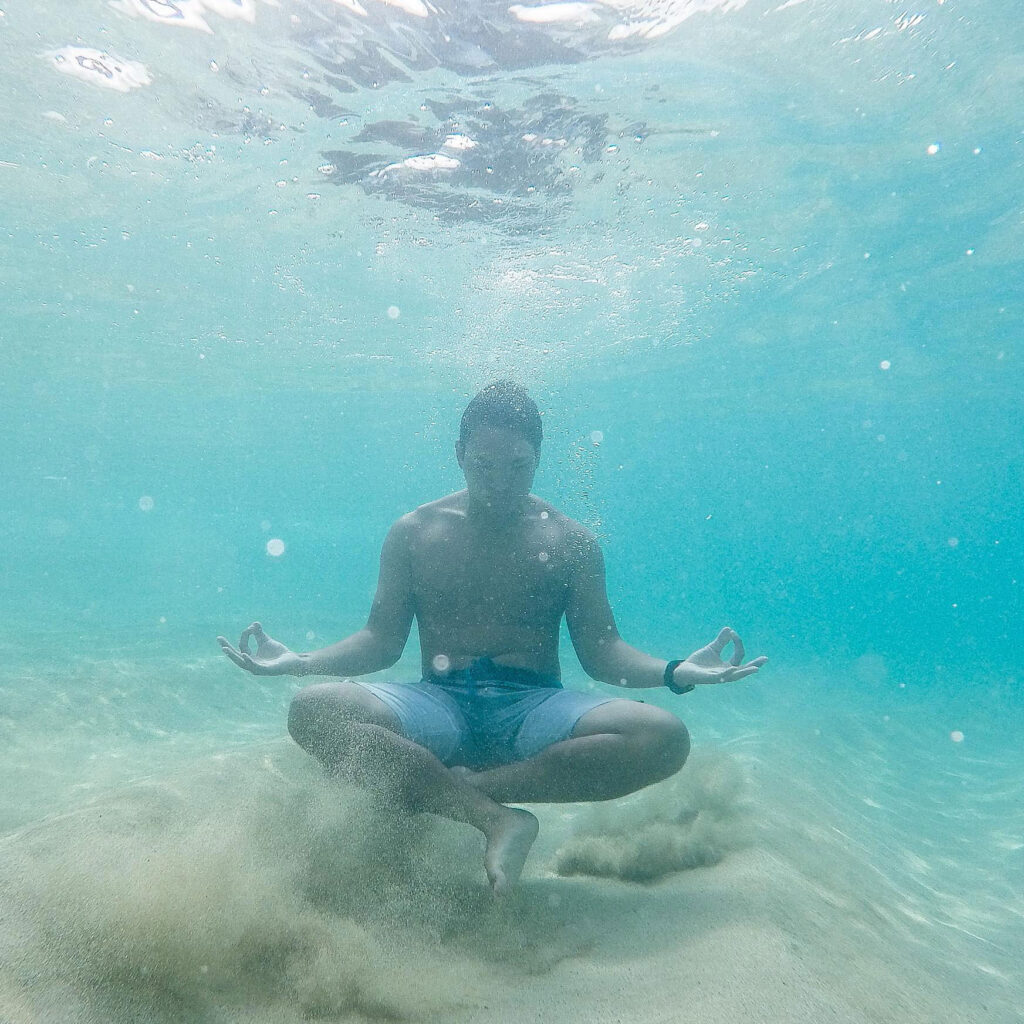 Self-Portrait of Raymond Enriquez under ocean water floating above sand with legs crossed and hands in a meditate stance, shirtless with blue shorts looking down toward clouds of sand.